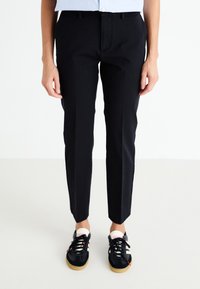Black tapered trousers with belt loops, front slant pockets, and a smooth texture, paired with black athletic shoes featuring white accents.