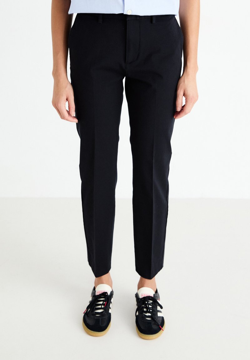 Black tapered trousers with belt loops, front slant pockets, and a smooth texture, paired with black athletic shoes featuring white accents.