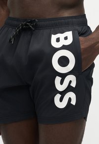 Mustat uimashortsit, joissa vasemman jalan valkoinen pystysuora "BOSS" logo sekä musta vyötärökiristys, jonka on solminut tumman ihon omaava henkilö.