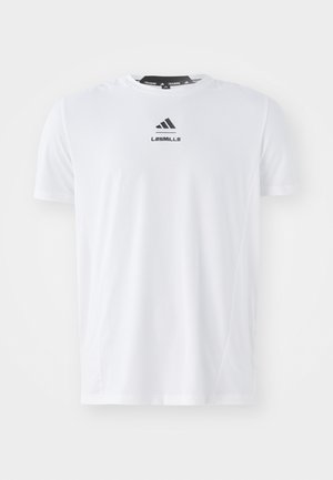 Camiseta atlética blanca fabricada en un material ligero. Presenta un escote redondo, mangas cortas y un logotipo negro de "LES MILLS" en la parte frontal.