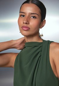 Grön ärmlös topp med draperat tyg och hög halsringning. Modellen har långt, rakt hår och bär ett guldörhänge i form av ett löv. Slät hudstruktur.