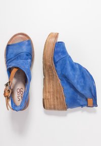 Pár modrých kožených wedges sandálů s dřevěnými podrážkami a opasky v tělové barvě, jedna bota z pohledu shora, druhá z boku.