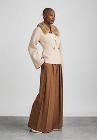 Beige cardigan med pelskrage og dobbelte knapper, kombinert med vide brune bukser. Teksturert stoff, skreddersydd design, og folder synlige.