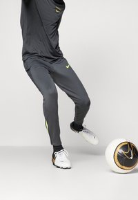 Grijze sportbroek met gele accenten, gecombineerd met witte voetbalschoenen en een zwart-wit gestreepte voetbals op een lichte achtergrond.