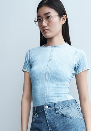 Νεαρή γυναίκα με μακριά σκουρόχρωμα μαλλιά που φορά διαφανή γυαλιά, ένα ανοιχτό γαλάζιο στενό τζιν πουκάμισο και ψηλόμεσα μπλε τζιν, μπροστά σε γκρι φόντο.