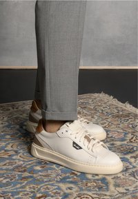 White leather sneakers with beige accents and a logo; round toe, flat sole, and textured detailing, paired with gray tailored trousers.
