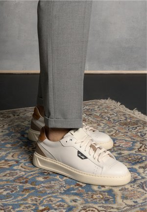 Baskets blanches en cuir avec des accents beiges et un logo ; bout rond, semelle plate et détails texturés, associées à un pantalon gris bien taillé.