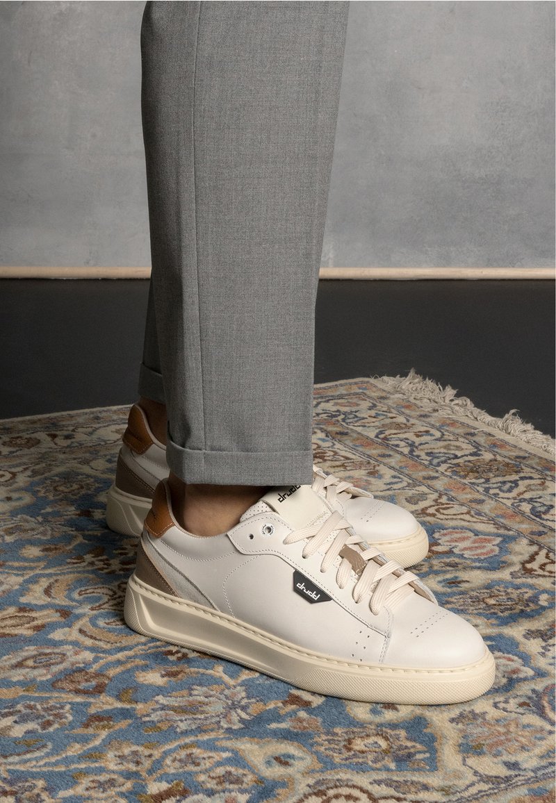 White leather sneakers with beige accents and a logo; round toe, flat sole, and textured detailing, paired with gray tailored trousers.