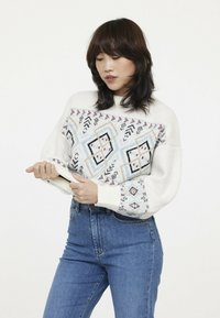 Pull crème avec des motifs géométriques en bleu, rouge et noir. Coupe ample avec des poignets et un col côtelés. Porté avec un jean bleu.