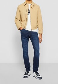 Beige bomber-takki vetoketjulla, valkoinen T-paita, jossa mustaa tekstiä, tummansiniset skinny-farkut ja mustavalkoiset tennarit.