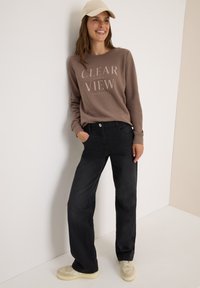 Sweatshirt marron avec le texte "CLEAR VIEW", jeans larges noirs et baskets beiges. Le sweatshirt a un col rond et des poignets côtelés.