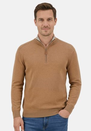 Man wearing a light brown zip-up sweater and blue jeans, standing with one hand in pocket, smiling slightly, against a plain light background.