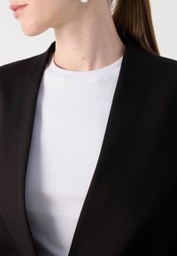 Blazer nero con un ampio risvolto, abbinato a una maglietta bianca a coste. Dettagli di orecchini argentati notevoli con una finitura liscia.