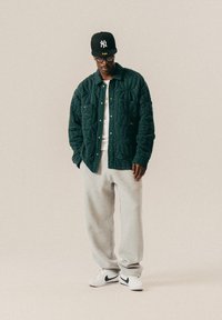 Άνδρας φορώντας ένα μαύρο καπέλο, γυαλιά, ραμμένη σκούρα πράσινη ζακέτα, λευκό πουκάμισο, γκρι φόρμες και λευκά αθλητικά παπούτσια με μαύρο λογότυπο στέκεται μπροστά σε απλό φόντο.