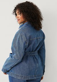 Denimjack in donkerblauw, met een riem in de taille, grote kraag en traditionele knoopsluiting. Zichtbare stiksels en ontspannen pasvorm.