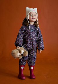 Dziecko w wodoodpornym kombinezonie w kwiatowy wzór i granatowym kolorze trzyma pluszową zabawkę, ma na sobie kremową czapkę w kształcie misia i burgundowe kalosze.