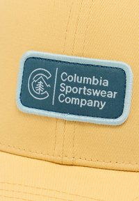 Κίτρινο καπέλο με μπλε ορθογώνιο ένθετο που φέρει το γράμμα «Columbia Sportswear Company» σε λευκό, παρουσιάζοντας υφασμάτινη υφή και δομημένο σχεδιασμό.