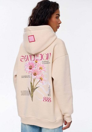 Beżowa bluza z kapturem z dużym różowym wzorem kwiatowym oraz napisem i akcentami, kwadratowa metka na kapturze i luźny krój.