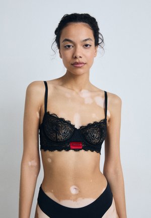 Black lace bra with underwire, scalloped edges, thin straps, and a central red logo tab; model in black briefs against a neutral background.