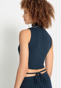 Frau mit lockigem Haar, die große Creolen und ein ärmelloses navyblaues Crop-Top mit gebundenen Trägern im unteren Rückenbereich trägt.