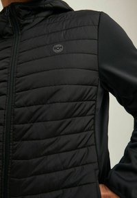 Chaqueta negra acolchada con cremallera frontal, textura acolchada y paneles laterales suaves. Incluye un pequeño logotipo circular en el pecho.