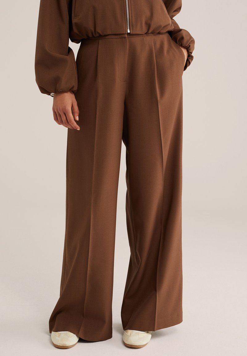 Personne portant un pantalon marron taille haute à jambes larges et des baskets blanches, avec une main dans la poche et l'autre relâchée sur le côté.