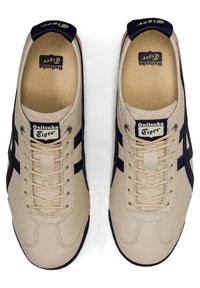 Beige leather sneakers with navy blue accents, featuring a lace-up design, rounded toe, and textured rubber sole. Brand logo on tongue.