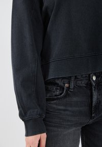 Felpa in cotone nera con polsini e orlo a coste, abbinata a jeans grigi scuri con chiusura a bottoni e una leggera trama.