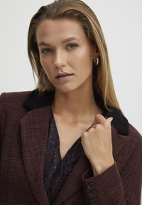 Blazer à motifs marron avec un col noir, doté de grands boutons, porté sur un haut floral sombre, mettant en avant un design structuré.