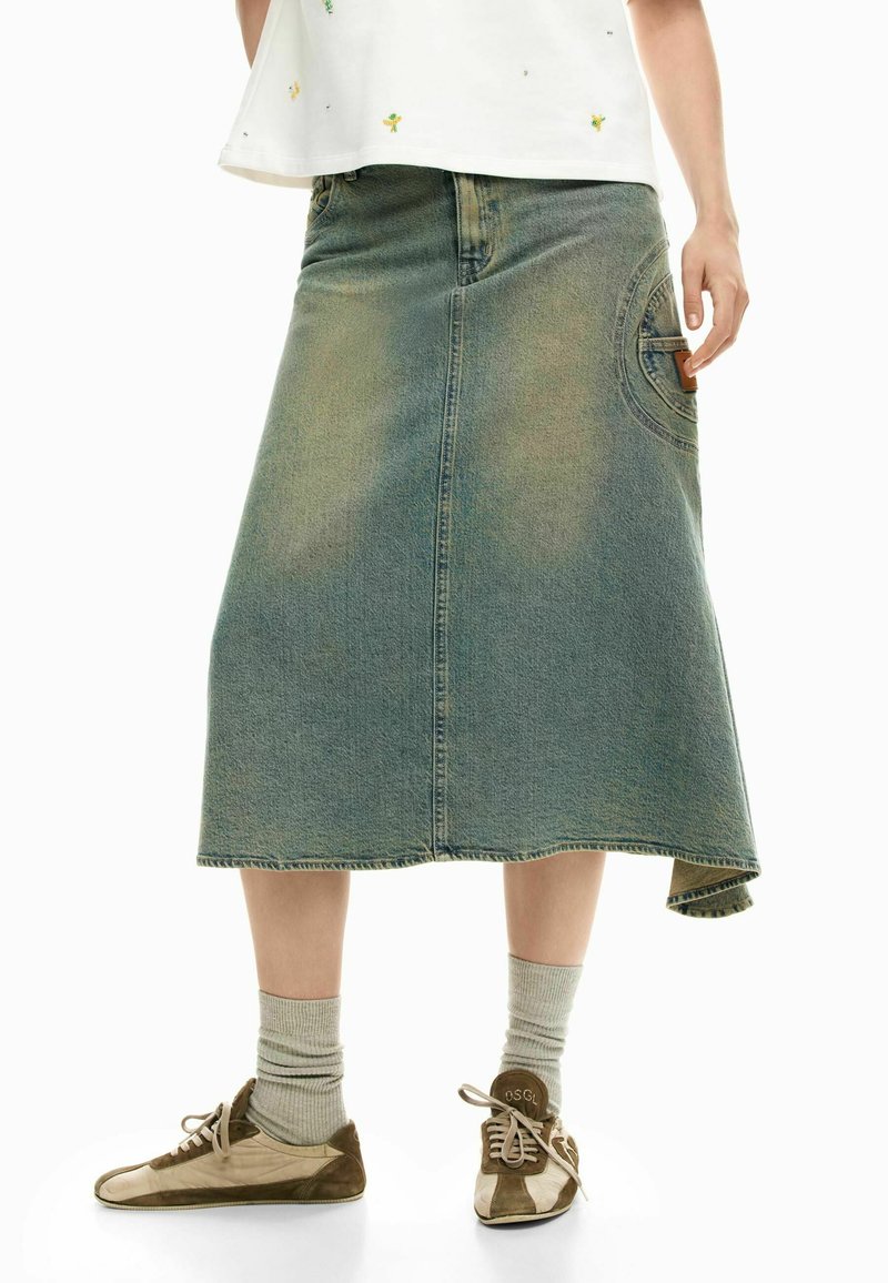 Persona che indossa una gonna midi in denim sbiadito, calzini grigio chiaro e sneaker marroni e beige con lacci, in piedi contro uno sfondo bianco.