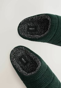 Pantofole in corduroy verde con soffice fodera in pelliccia sintetica grigia, dotate di una soletta etichettata. Forma curva con texture rigata e parte superiore imbottita.