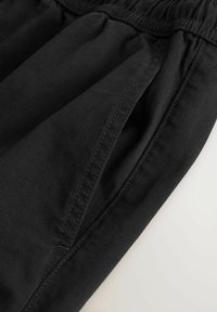 Gros plan sur un pantalon en tissu noir avec une ceinture élastique et une poche latérale visible sur un fond clair.