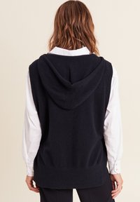 Un hoodie sans manches en tricot navy avec une finition texturée, doté d'une capuche spacieuse et d'une finition côtelée, porté sur une chemise blanche à col.
