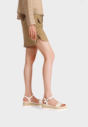 Donna che indossa pantaloncini beige, un maglione a maglia e sandali bianchi con zeppa e cinturini intrecciati, in piedi di lato su uno sfondo semplice.