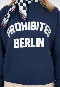 Sweat-shirt bleu marine avec texte blanc "PROHIBITED BERLIN" et une écharpe à carreaux bleu marine, blanc et rouge autour du cou.