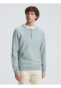 Pulóver de punto de manga larga azul claro con un cuello henley abotonado, puños y dobladillo acanalados; usado sobre una camiseta blanca.