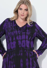 Schwarzes Tunika-Kleid mit lila Batikmuster, V-Ausschnitt, langen Ärmeln, seitlichen Taschen und weichem Stoff, der für Komfort sorgt.