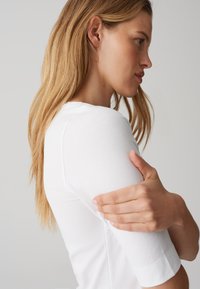 Frau mit langen blonden Haaren in weißem Kurzarmshirt hält ihren Oberarm, blickt nach rechts vor neutralem grauem Hintergrund.