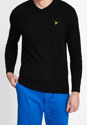 Homme portant un pull noir à manches longues en V avec un petit logo d'oiseau jaune et un pantalon bleu vif, main dans la poche.
