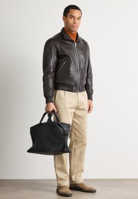 Giubbotto bomber in pelle marrone, pantaloni beige e stivali in suede marrone. Tiene una borsa nera con texture, manici doppi e tracolla regolabile.