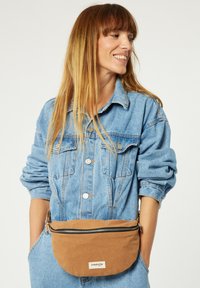 Sac banane en tissu beige avec une fermeture éclair, porté sur une veste en jean bleu clair. Présente une étiquette de logo simple et une forme circulaire et douce.