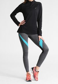 Černé tričko s dlouhým rukávem a zipovým límcem, šedé legíny s modrými akcenty a černými panely, doplněné růžovými a šedými sportovními botami.