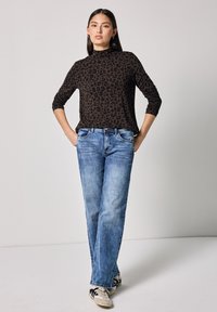 Braun gepunktete Langarmbluse kombiniert mit hellblauen geraden Jeans und cremefarbenen Sneakers. Einfaches Design, weicher Stoff und lässiger Stil.