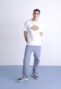 Valkoinen t-paita, jossa on pyöreä Dickies-logotulostus, yhdistettynä vaaleanharmaisiin housuihin ja harmaisiin urheilukenkiin. Tausta on yksinkertainen ja vaalea.