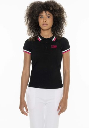 Femme aux cheveux bouclés portant un polo noir avec des bordures roses et blanches et un pantalon blanc, debout devant un fond blanc.