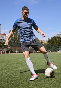 Camisa deportiva gris con acentos azules y logotipo, pantalones cortos negros, calcetas blancas y zapatos de fútbol con acentos rojos. Jugador interactuando con un balón de fútbol.