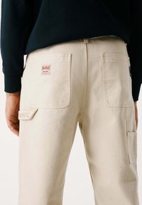 Beige Cargo-Hosen aus strapazierfähiger Baumwolle mit zwei Gesäßtaschen, ausgestattet mit einem Red Bull Racing Logo-Patch und gestickten Details.