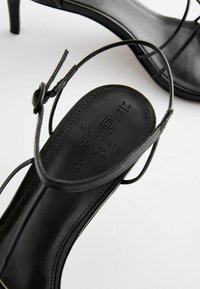 Svarta klacksandaler med tunna remmar, med en slät lädersula präglad med varumärkesdetaljer och storleksmarkering.
