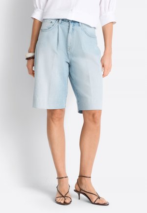 Frau trägt hellblaue Denim-Bermudashorts, weiße Bluse, braune Sandaletten mit Absatz und Riemchen sowie Armreifen am linken Handgelenk, steht vor weißem Hintergrund.