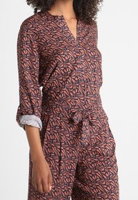Tuta alla moda con maniche lunghe e colletto, caratterizzata da un motivo geometrico nei colori rosso, blu navy e crema; il materiale è leggero, con una vita regolata da una cintura e dettagli con risvolto sulle maniche.